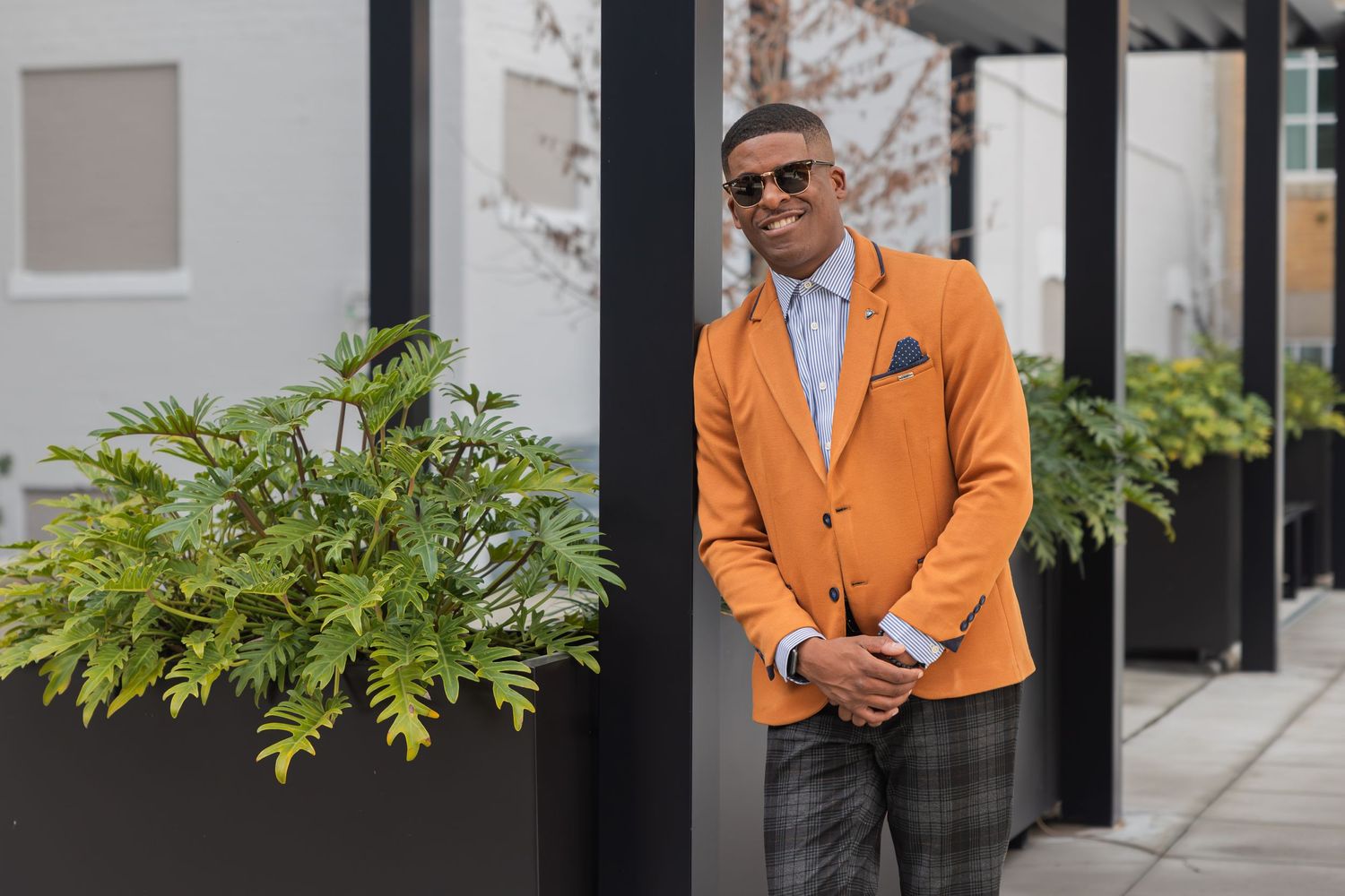 Man dressed in orange suit jacket with sunglasses smiling leaning against a pole