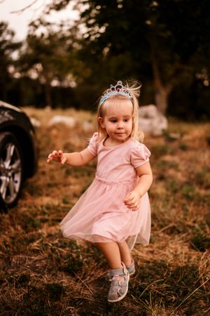 Young child in pink dress and tiara dances playfully outdoors.