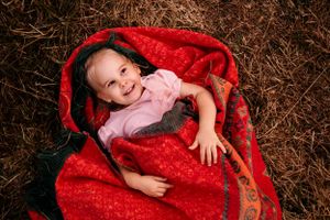 Child wrapped in a cozy red blanket lying on hay, smiling during an outdoor photo session.