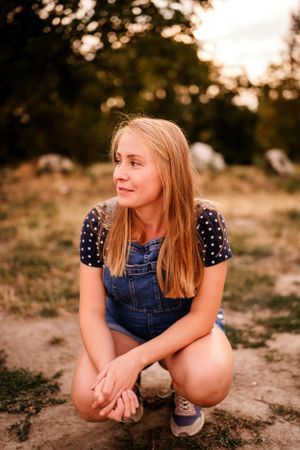 Woman in polka dot top and denim overalls crouches outdoors at sunset.