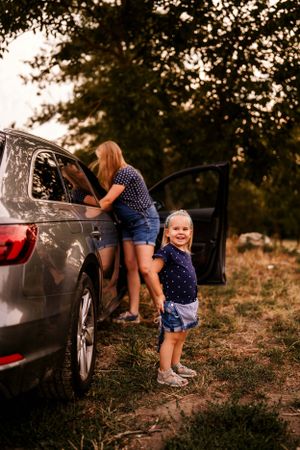 A heartwarming scene by a car as one stands in denim overalls while another poses near the door.