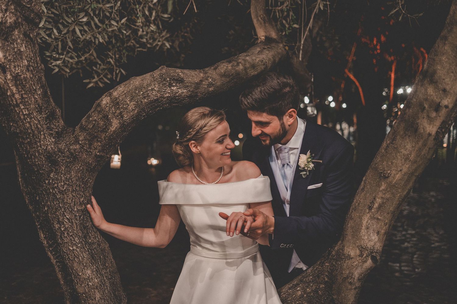 Newlyweds sharing intimate moments under a canopy of trees with twinkle lights at dusk.