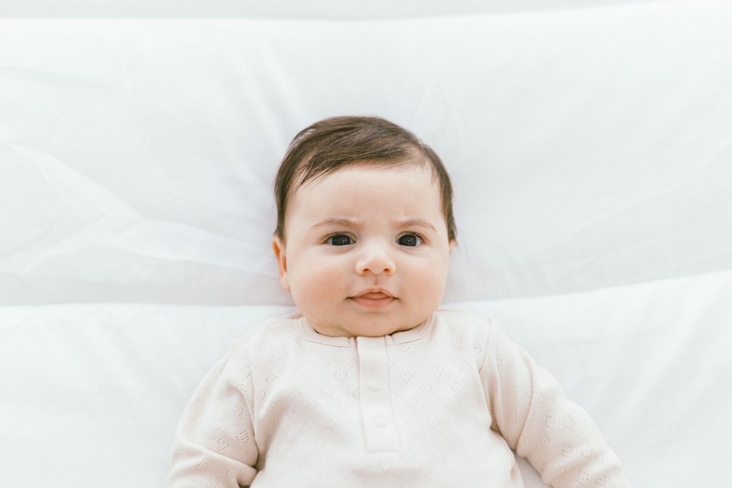 Baby in light beige outfit lying on white bedding with sweet smile.