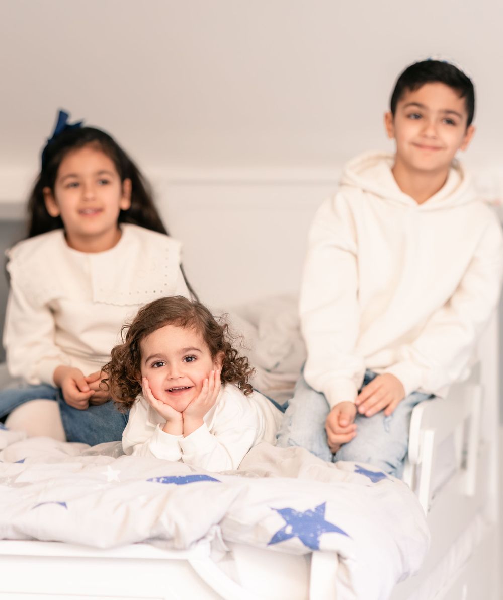 Three children in white clothing sitting casually on a bed with star-patterned bedding.