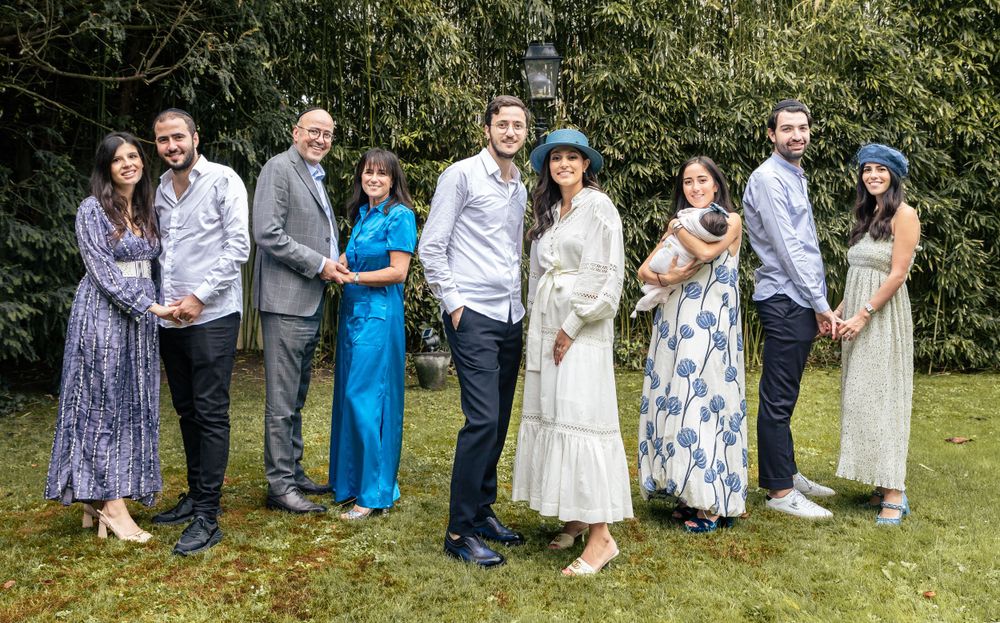 Wedding party in formal attire poses together outdoors with greenery backdrop.