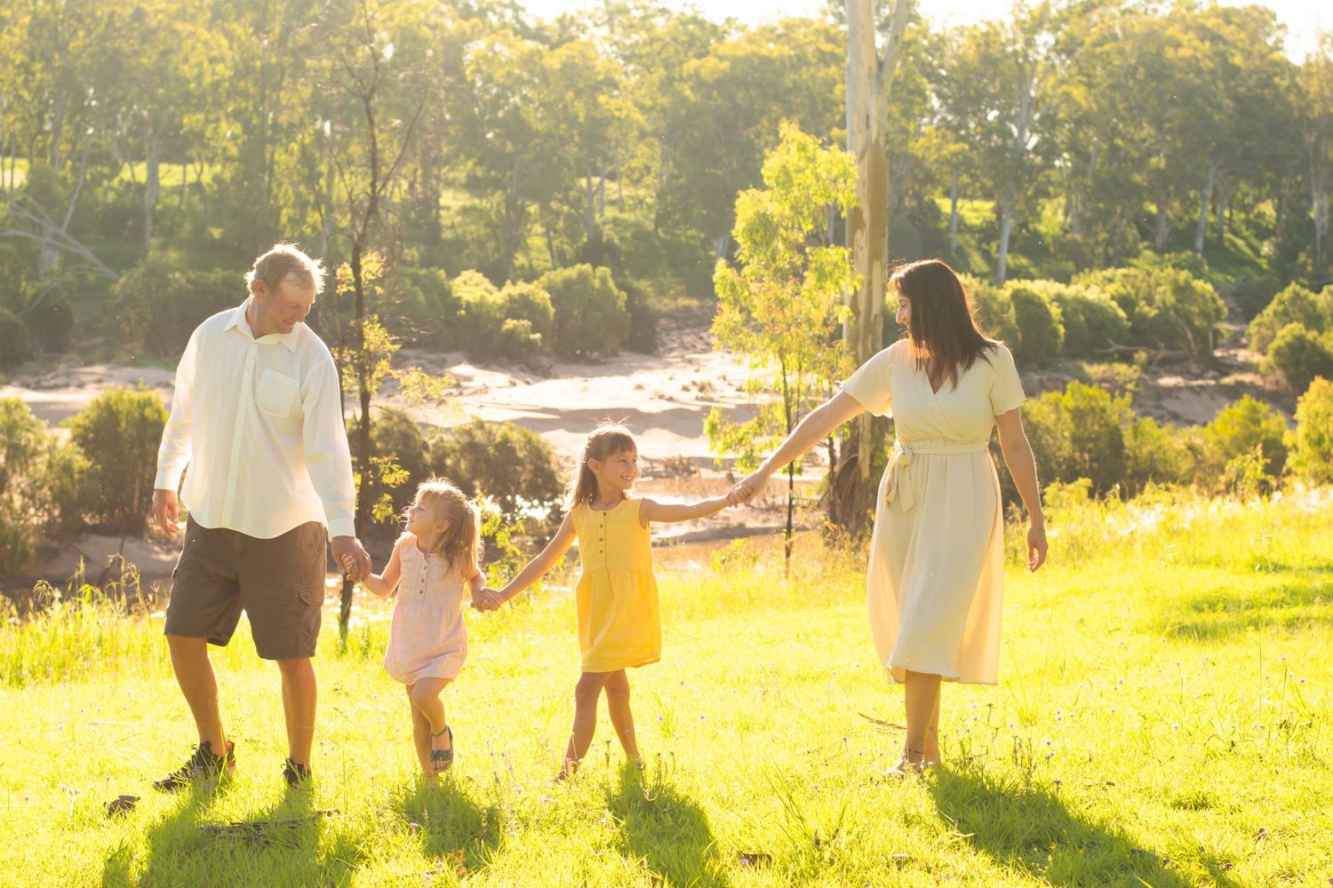 Family of four holding hands while walking through sunlit field at golden hour.