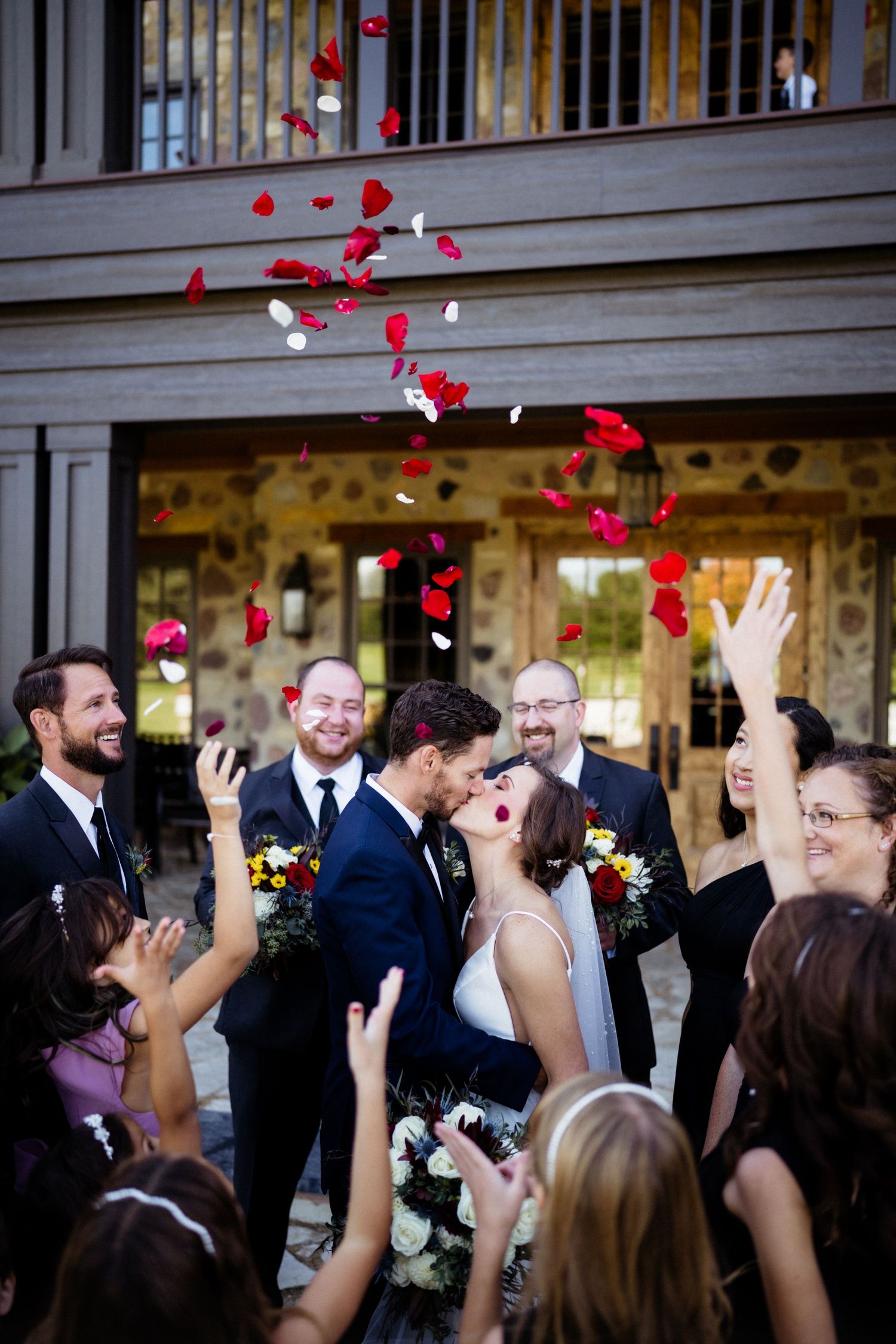 A joyous wedding celebration with red rose petals being tossed in the air outside a stone building.
