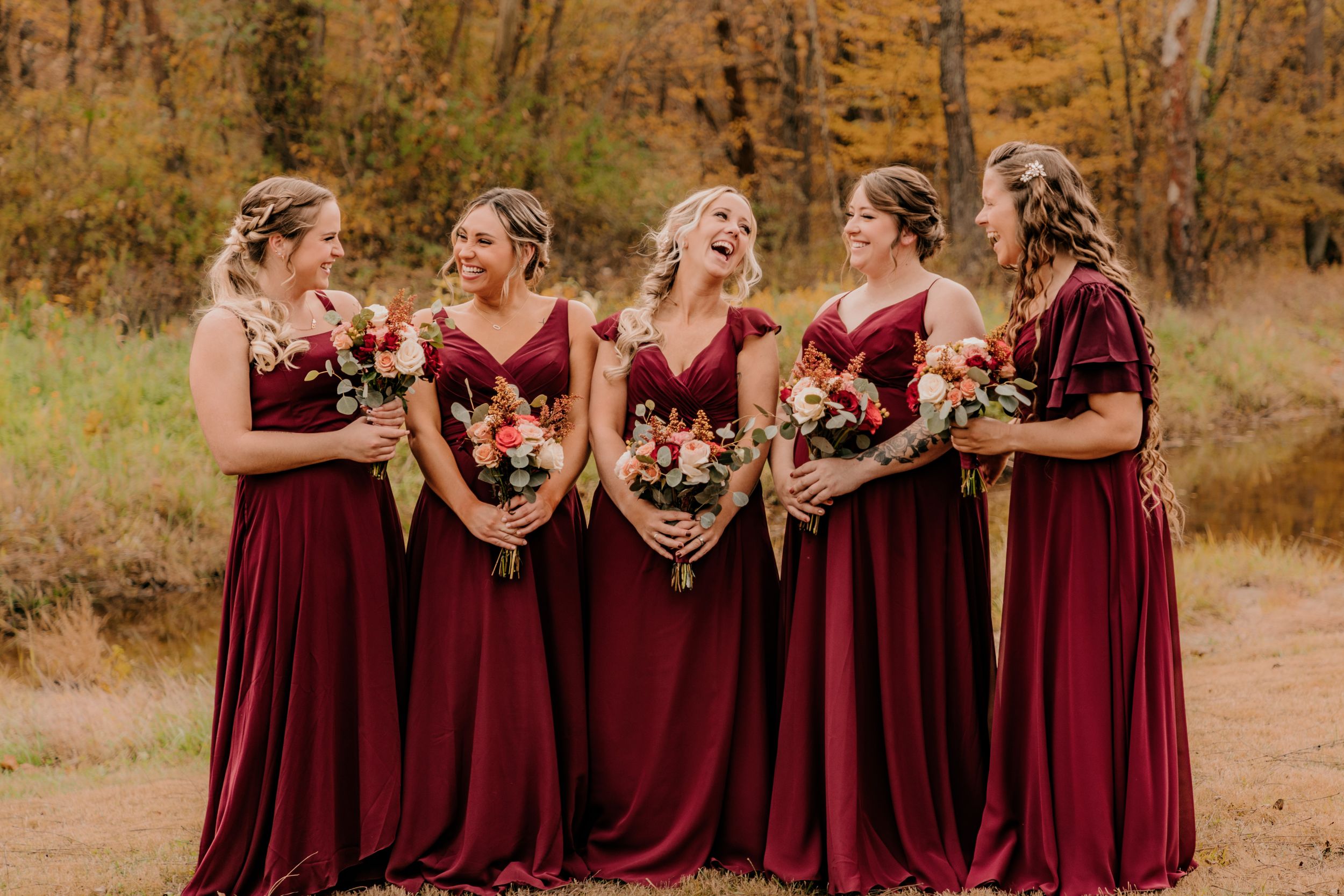 Bridesmaids in burgundy dresses holding bouquets against fall foliage.