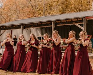 Row of bridesmaids in burgundy dresses standing in autumn forest setting.