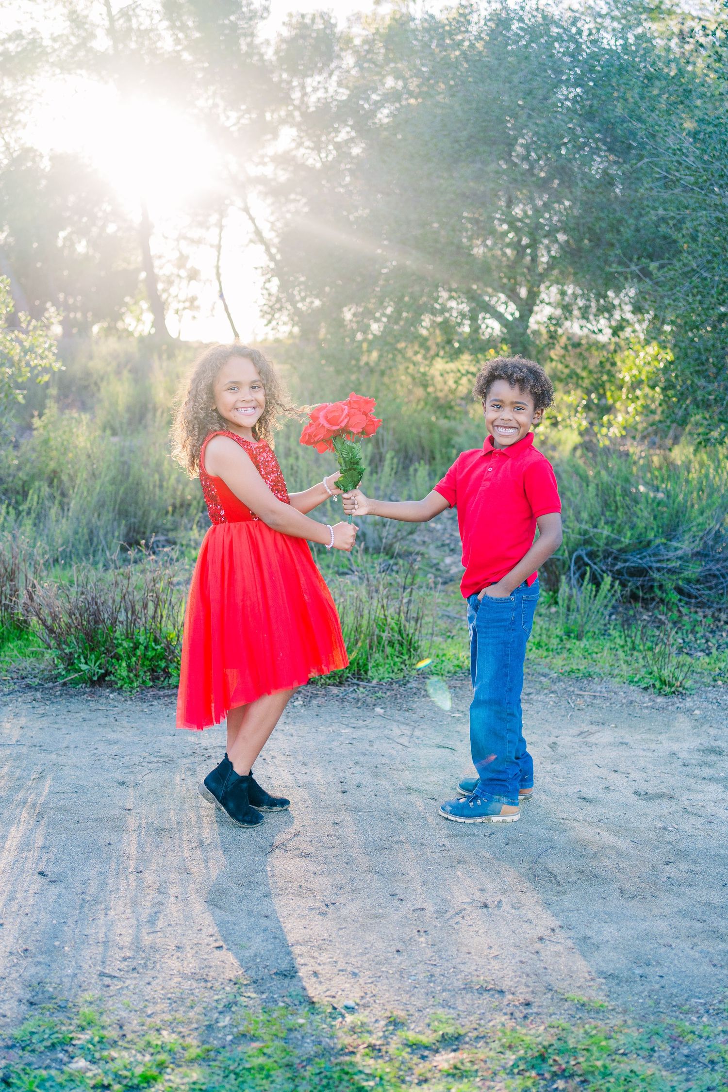 Two children stand on a dirt path in nature, wearing red and exchanging flowers in golden sunlight.