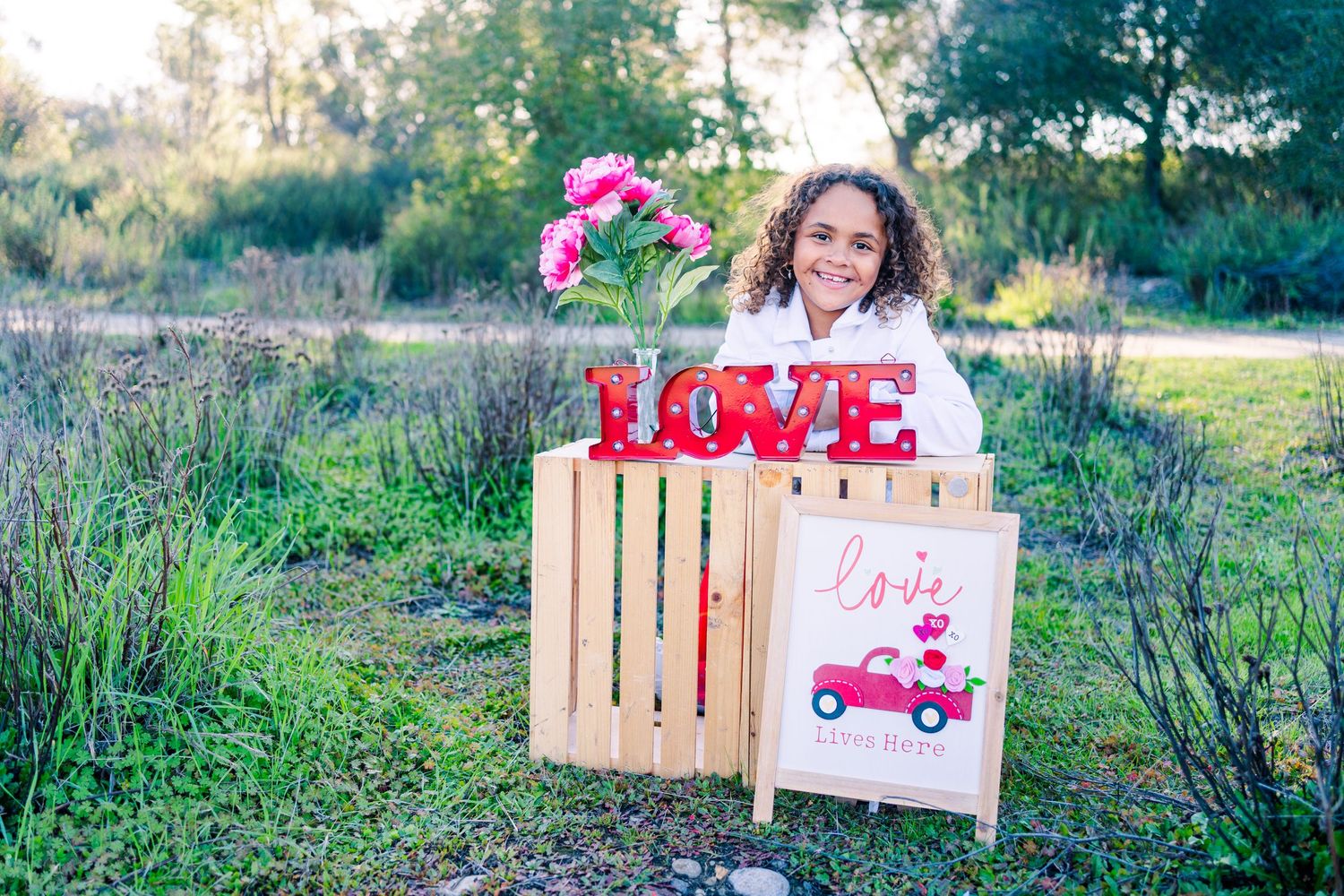 Valentine's Day photo setup in nature with red LOVE letters, wooden crate, vintage truck sign and pink flowers.