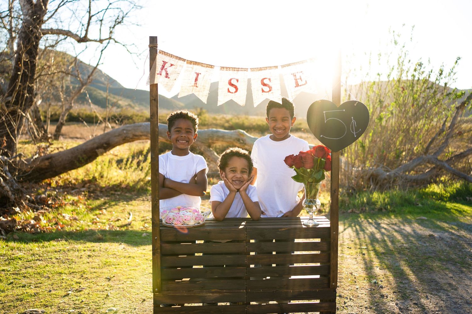 Children at rustic wood kissing booth with heart decorations during golden hour in outdoor garden setting.