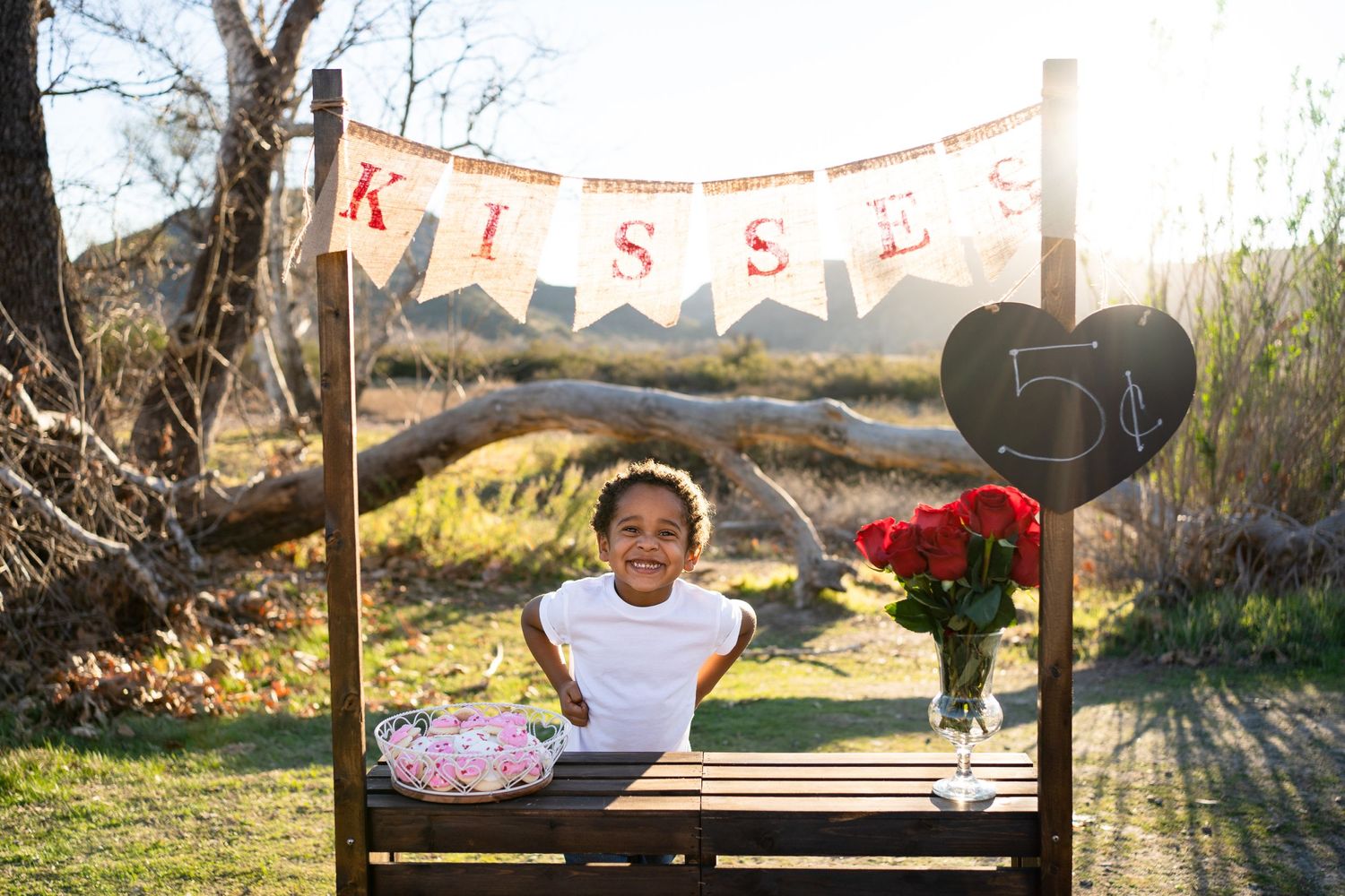 A wooden kissing booth display set up in a sunny garden with roses and rustic banners for Valentine's Day.
