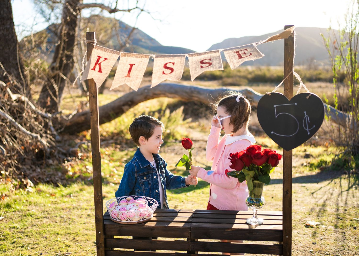 Two children sit at an outdoor kissing booth with red roses and heart decorations on a sunny day.
