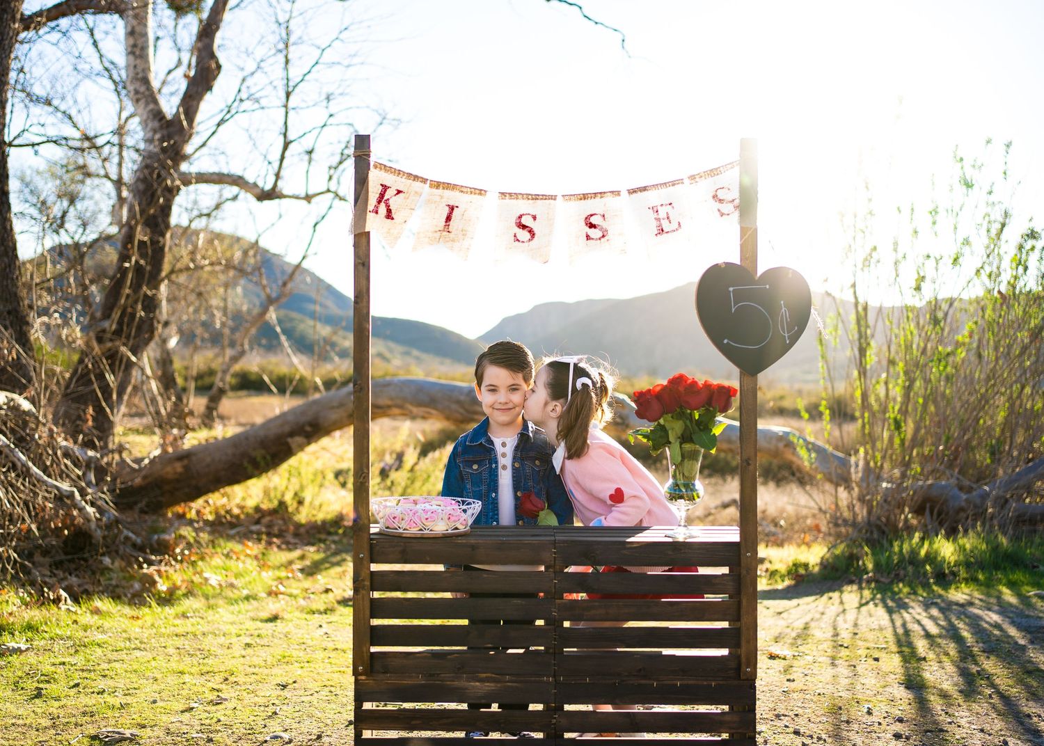 Romantic outdoor kiss booth decorated with banner and heart balloons against scenic mountain backdrop at sunset.