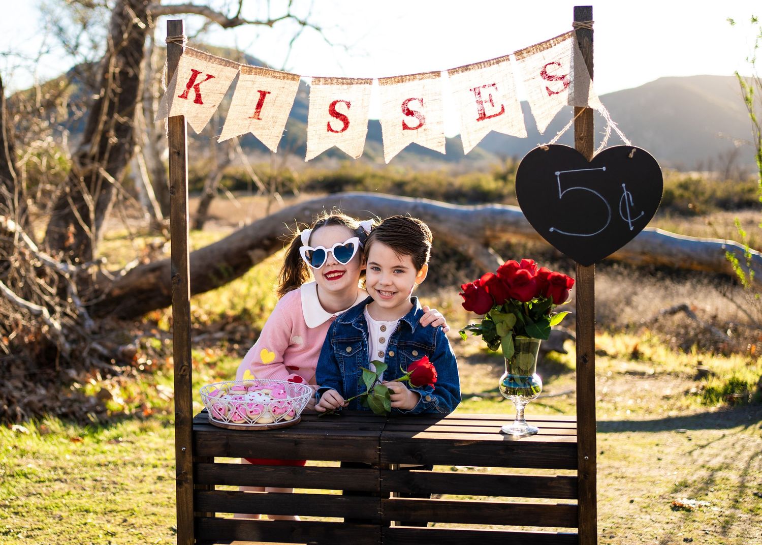 A rustic wooden bench with a KISSES banner and romantic valentine themed decor in a scenic outdoor setting at sunset.