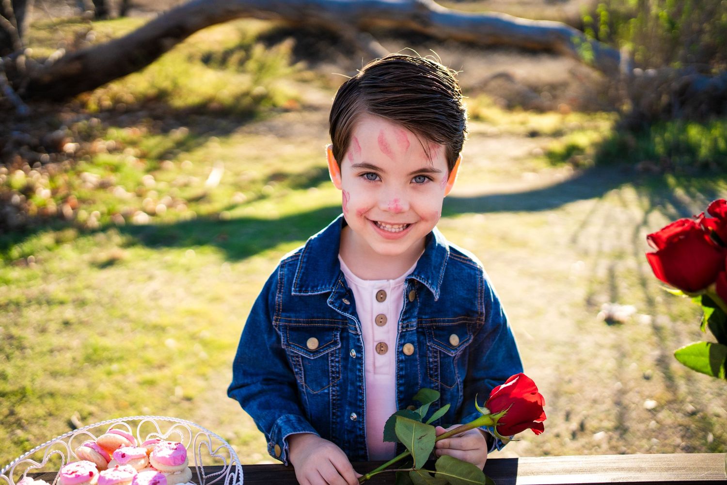 Young person in denim jacket poses outdoors holding red roses with blurred nature background.