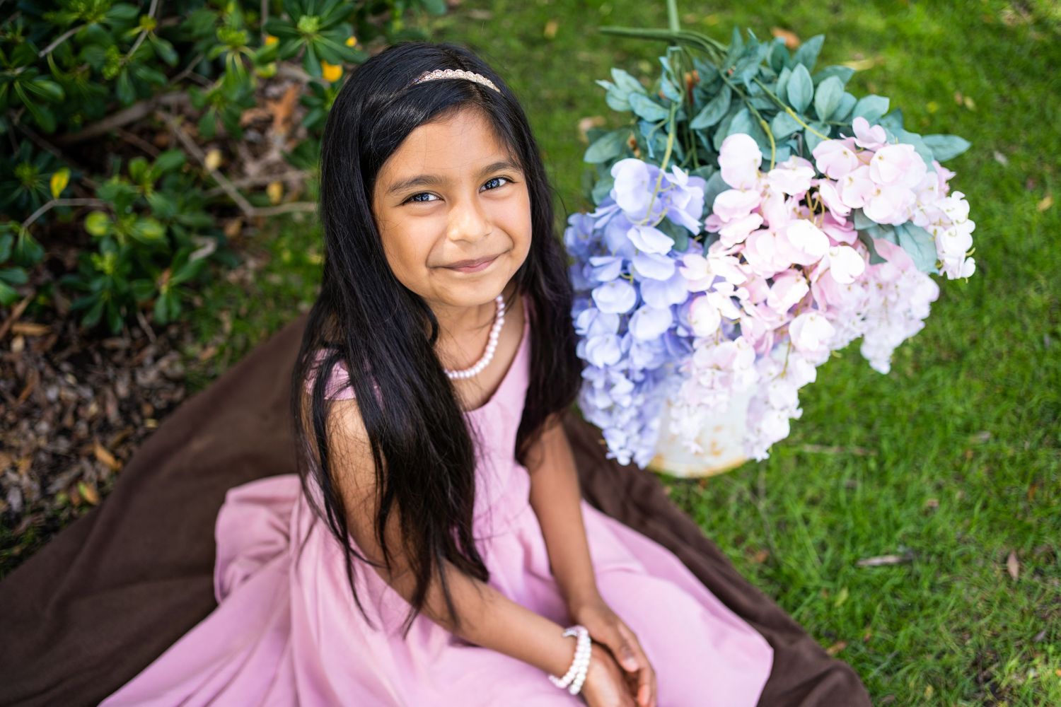 Someone in a pink dress sits on a brown blanket on grass with purple and pink hydrangeas in the background.