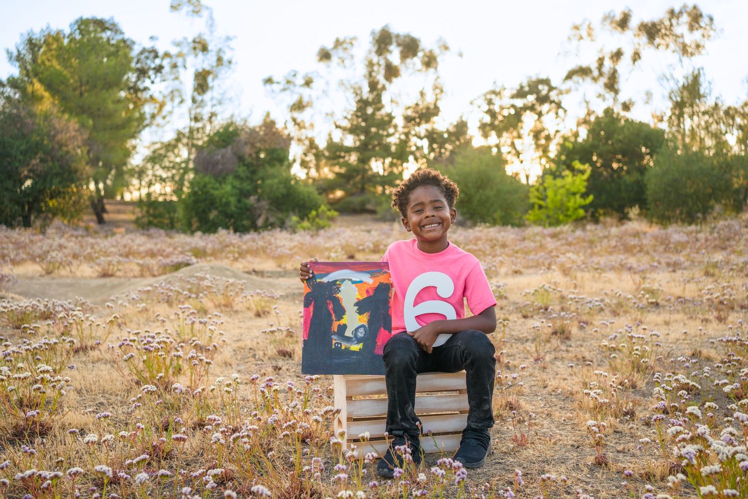 Person in a pink shirt sits on a wooden crate in a golden field during sunset, holding artwork.