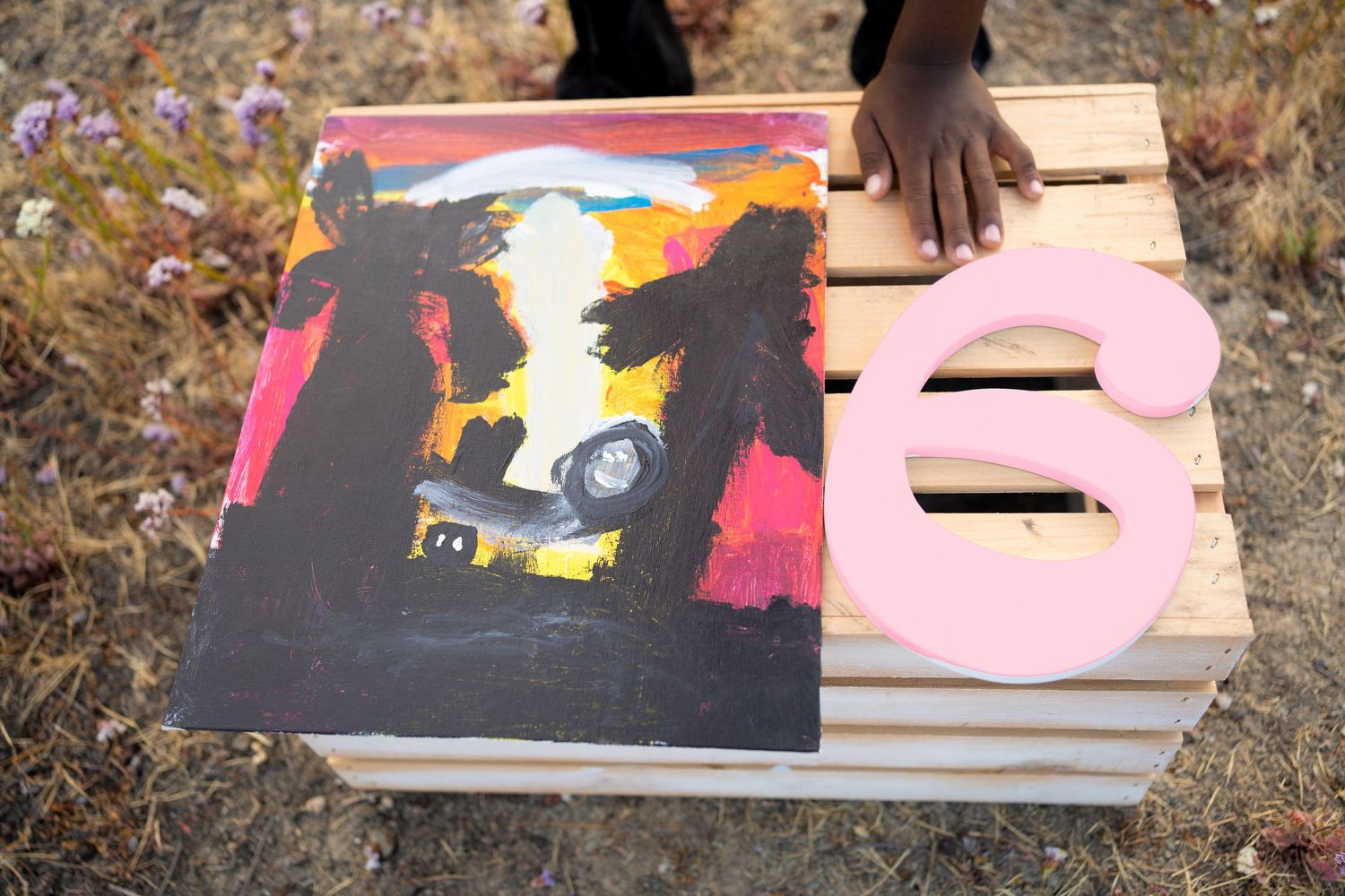 Artistic painting on black canvas displayed outdoors with a pink decorative element on wooden surface.