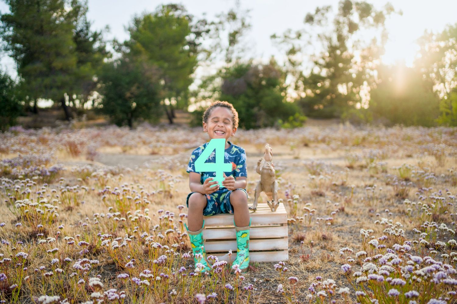 A small reader sits on stacked books in a field of wildflowers at sunset while wearing teal colored accessories.