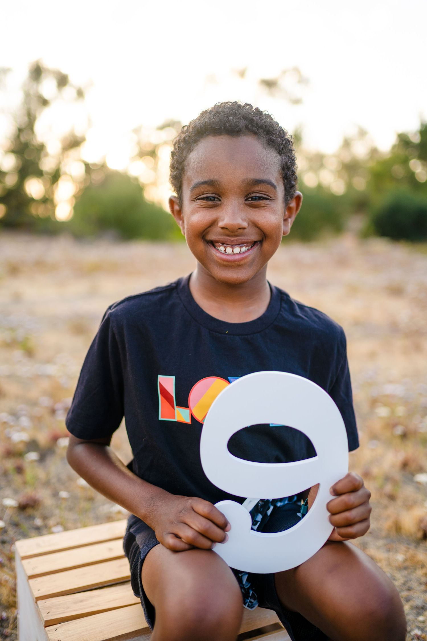A smiling youth in a black t-shirt holds a large white number 9 while sitting on a wooden chair at sunset.