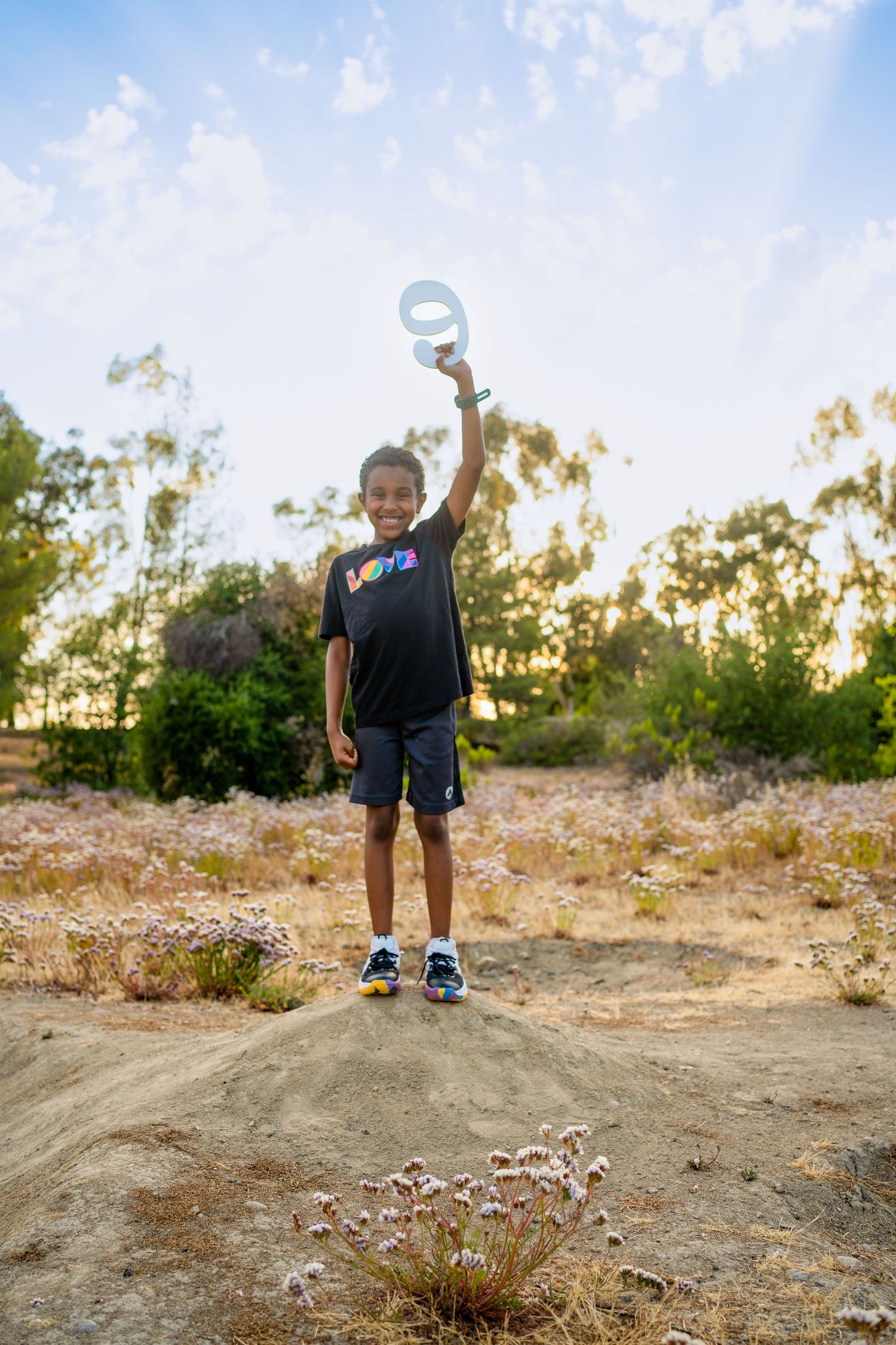 Young child stands triumphantly on dirt mound with arms raised during sunset in outdoor nature setting.