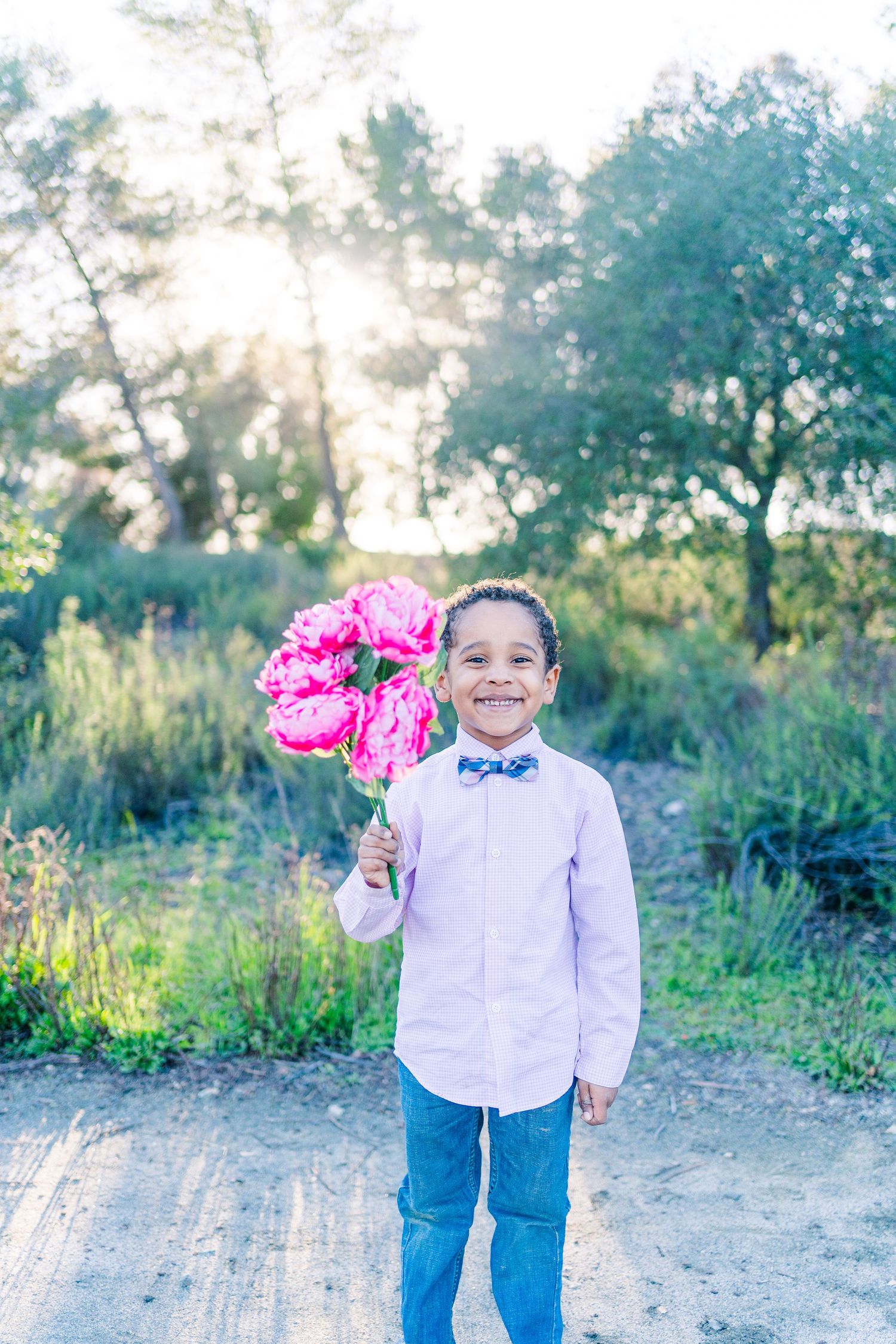 A young figure in a blue bowtie and white shirt holds bright pink flowers on a sunny outdoor path.