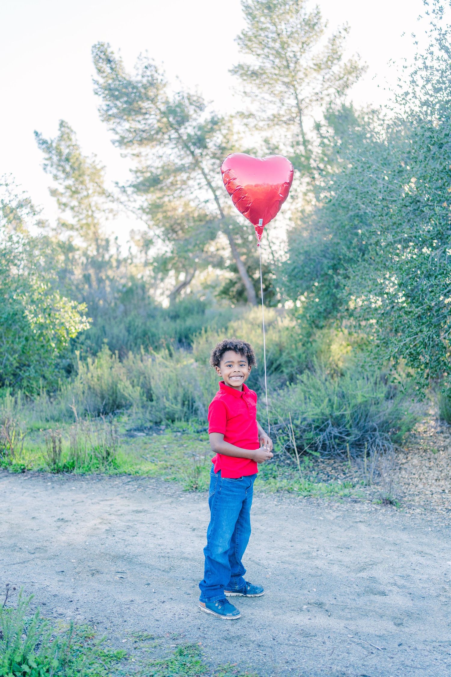 A red heart-shaped balloon floats above a path surrounded by green trees and shrubs in a serene outdoor setting.