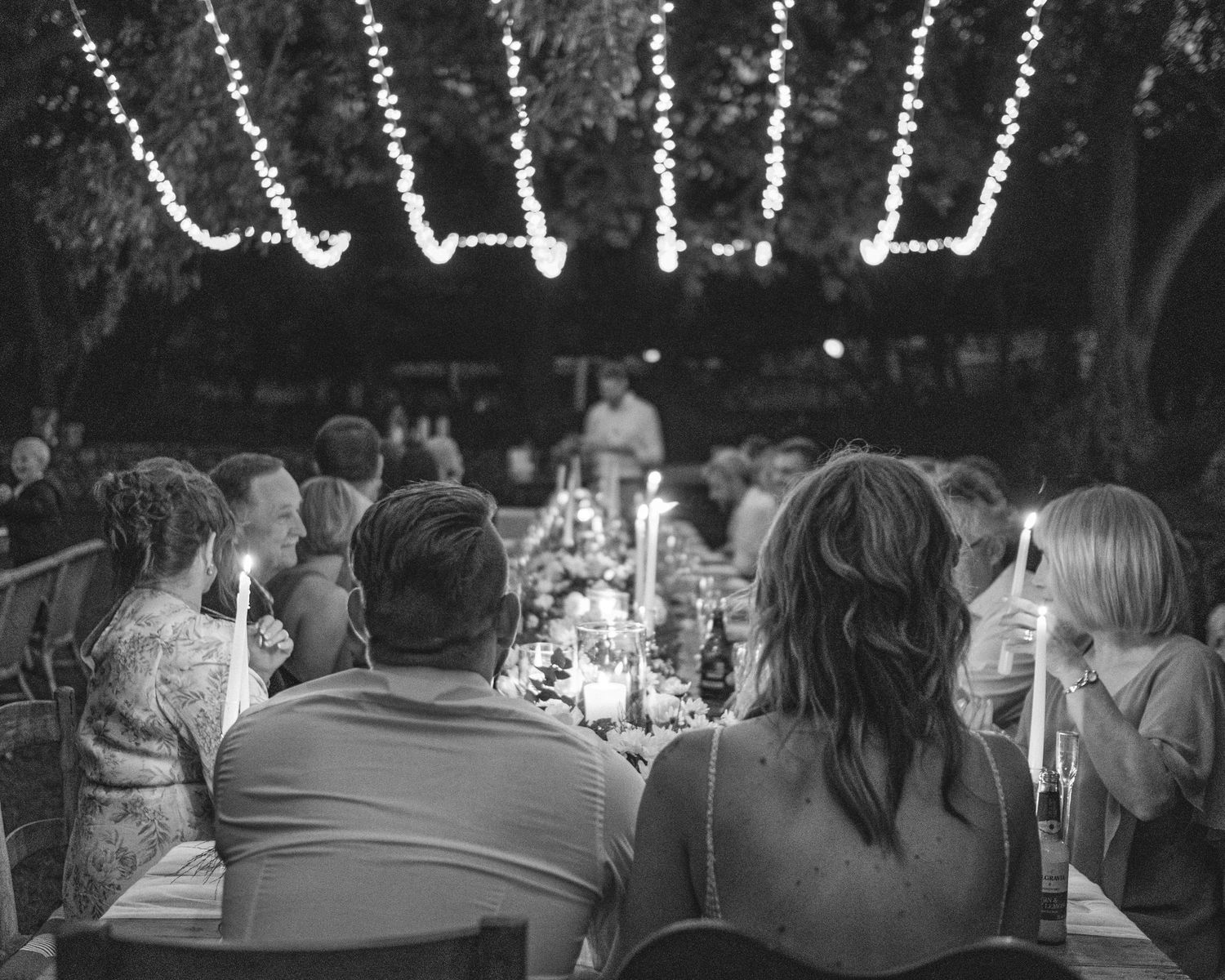 Bride and groom sitting at end of table under fairy lights during outside reception during wedding at Intiem Weddings, c