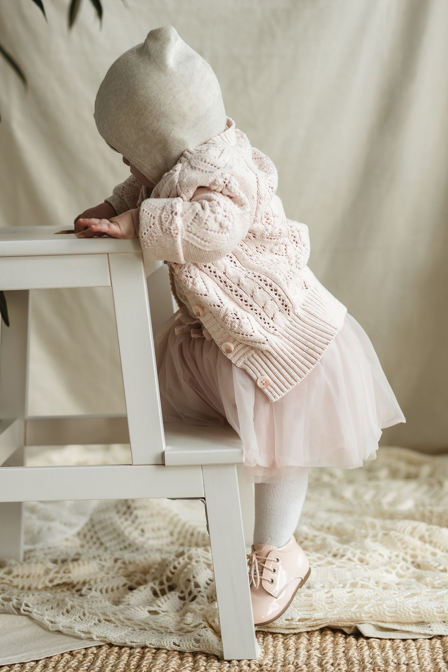 Back view of toddler in pink dress and white bonnet leaning on white chair.