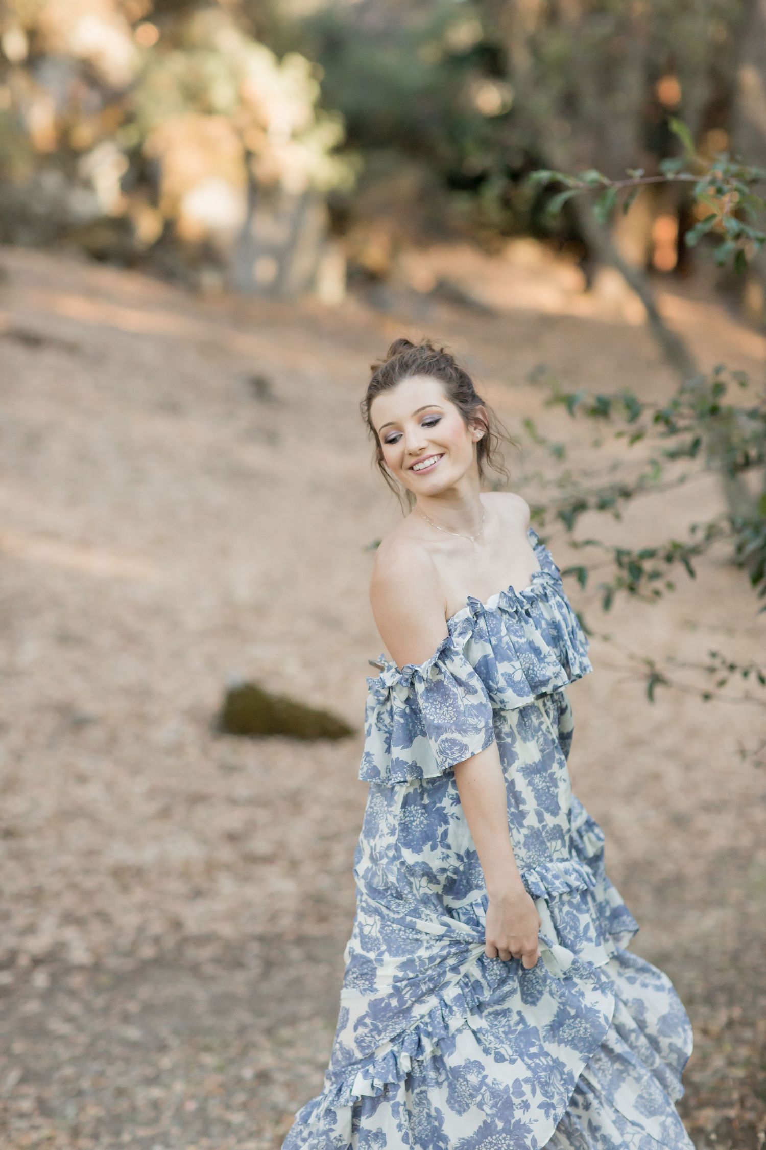 Joyful young woman twirling in a blue floral off-shoulder dress in a sunlit outdoor woodland setting.
