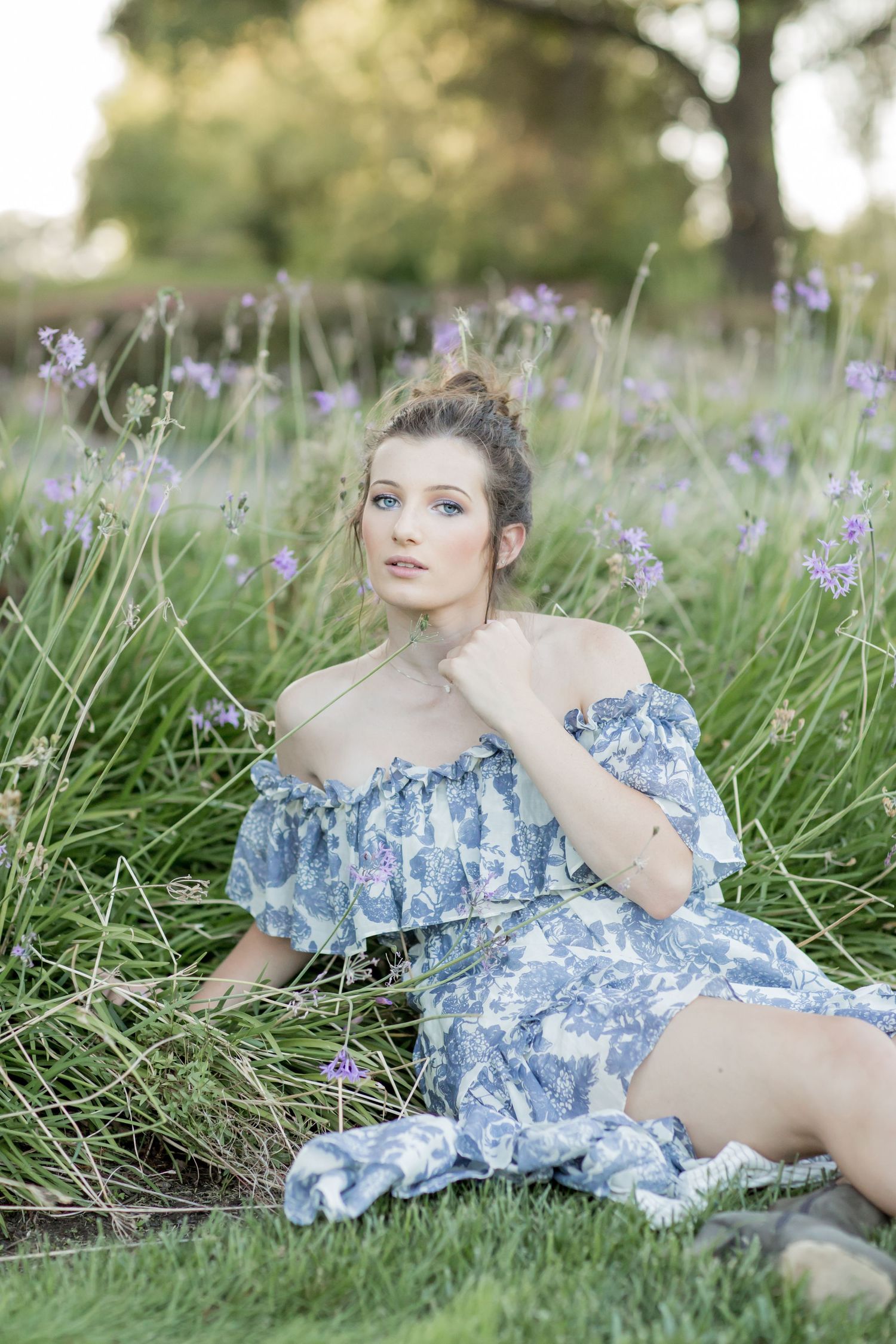 Young woman in blue floral off-shoulder dress sitting gracefully among tall purple wildflowers outdoors.