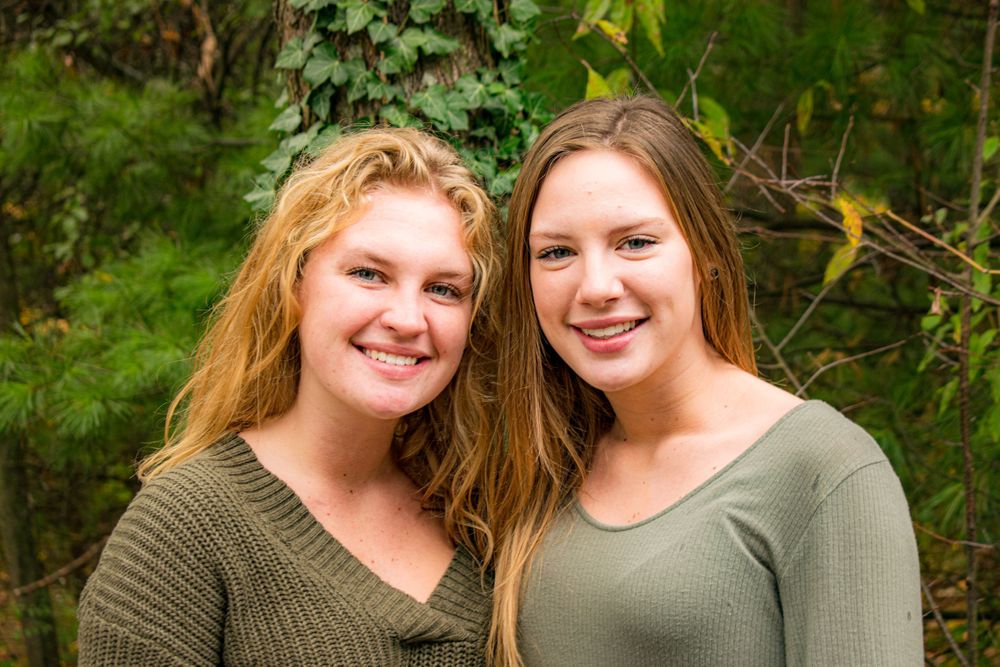 Two women in olive green tops pose outdoors against a natural green background.