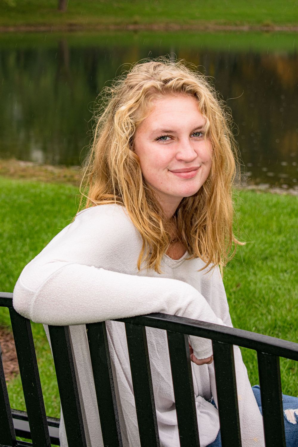 Person in a white sweater sitting on a park bench with messy blonde hair on a sunny day.