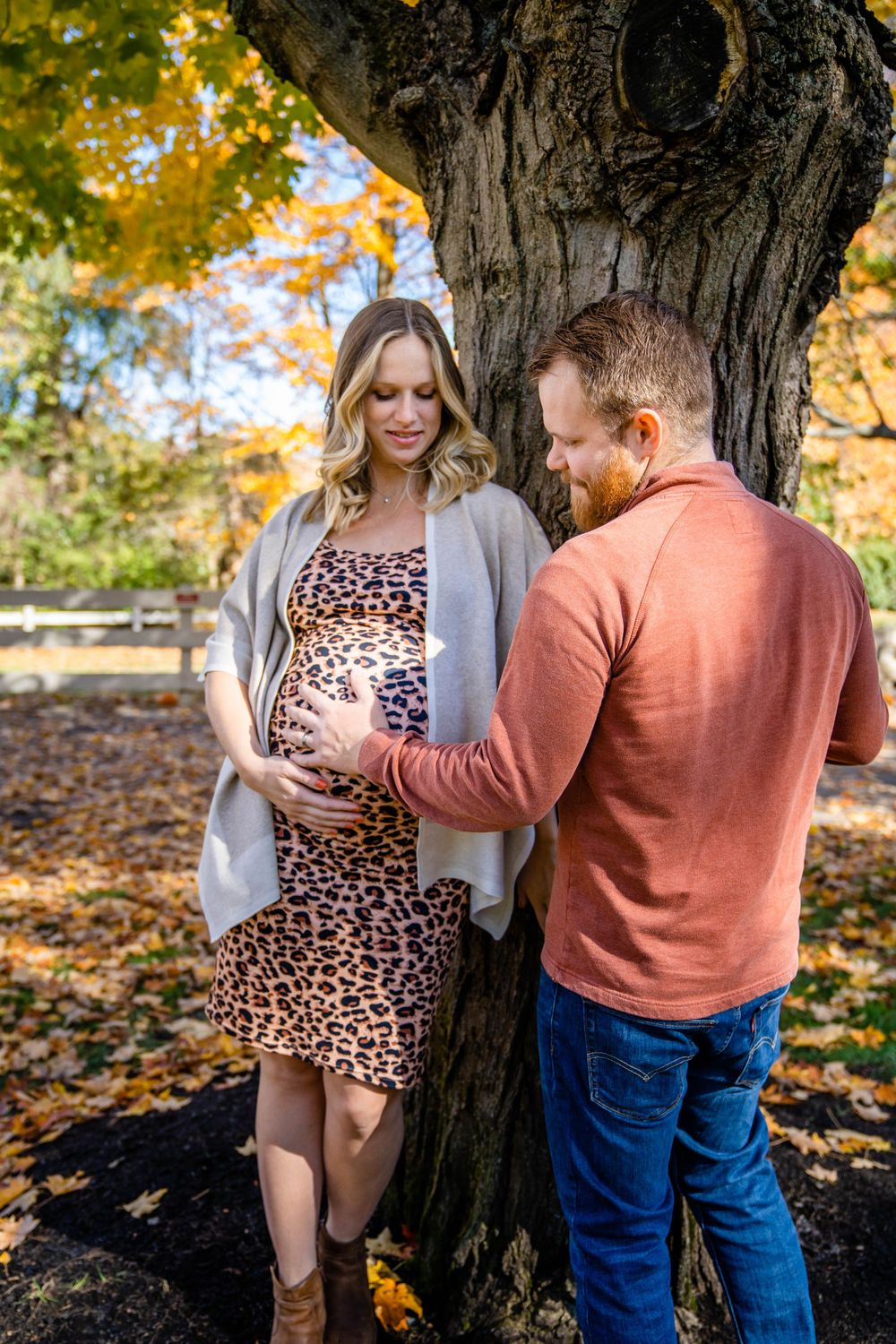 Autumn maternity photo of couple standing by large tree trunk with golden leaves above.