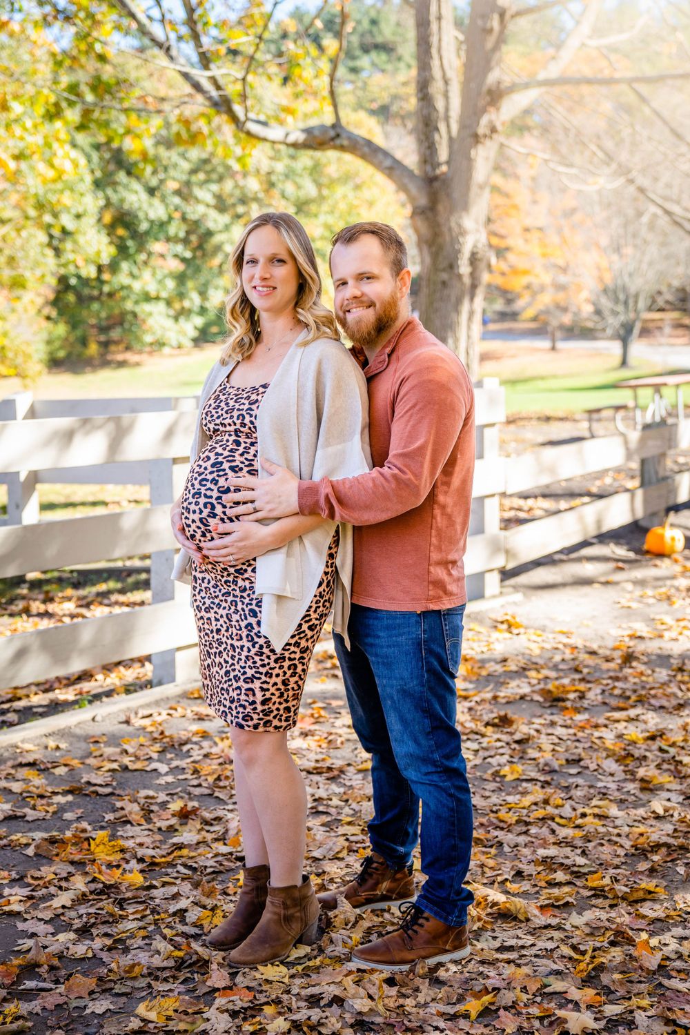 Expectant couple poses by wooden fence surrounded by autumn foliage in outdoor maternity photoshoot.