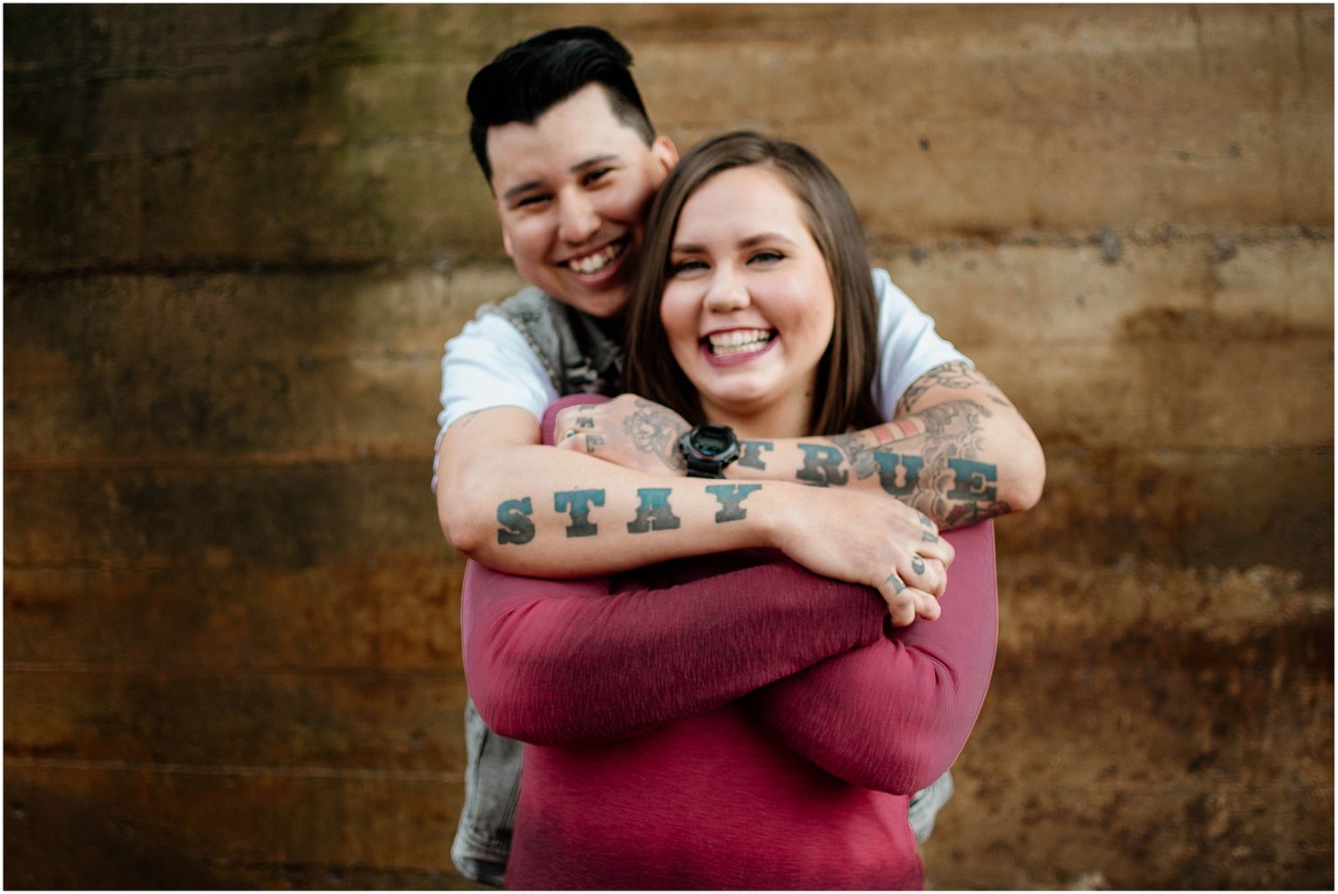 Couple shares loving embrace while showcasing visible tattoos against rustic wall background.