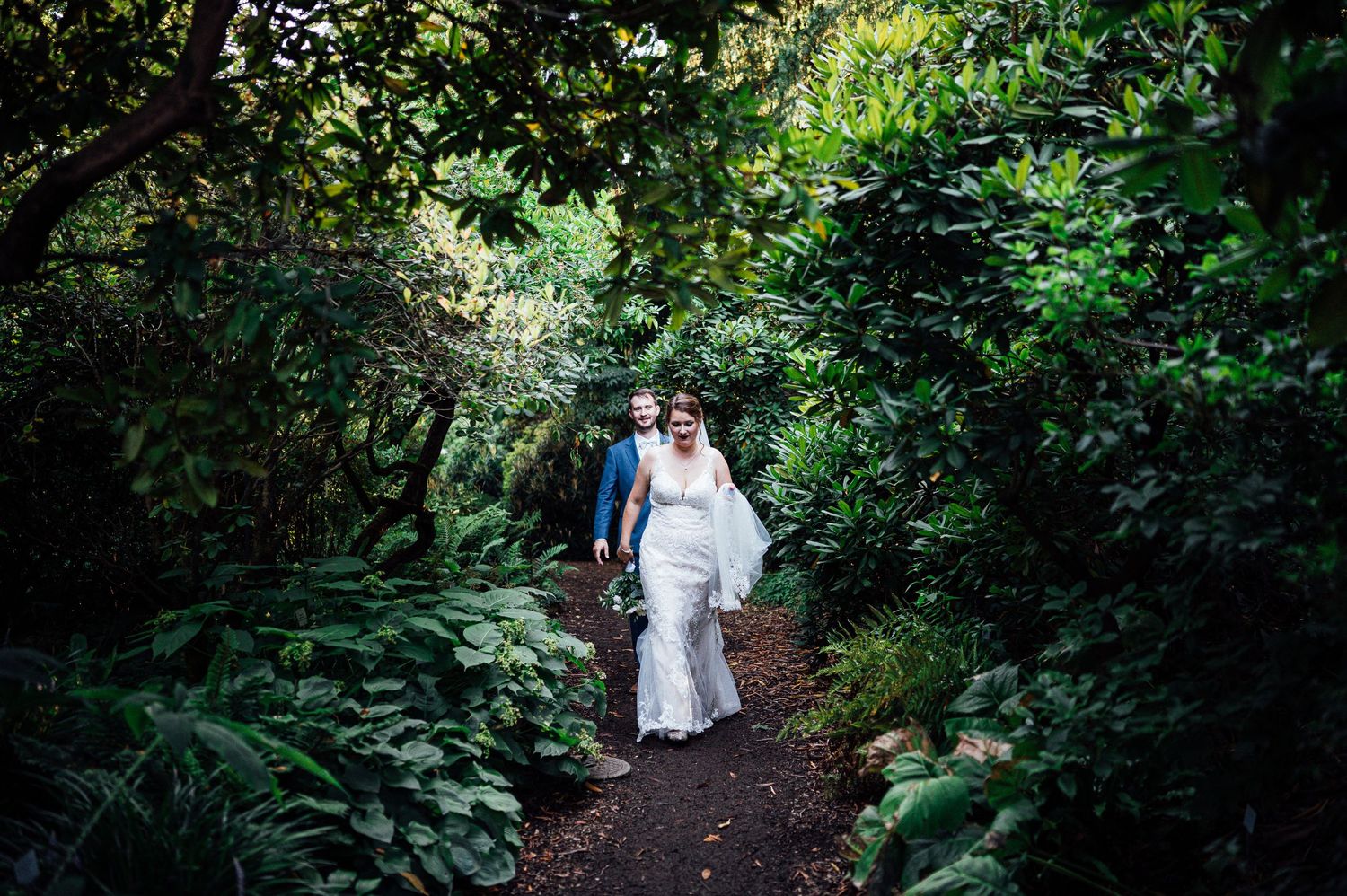 Couple walking along lush green forest path in wedding attire.