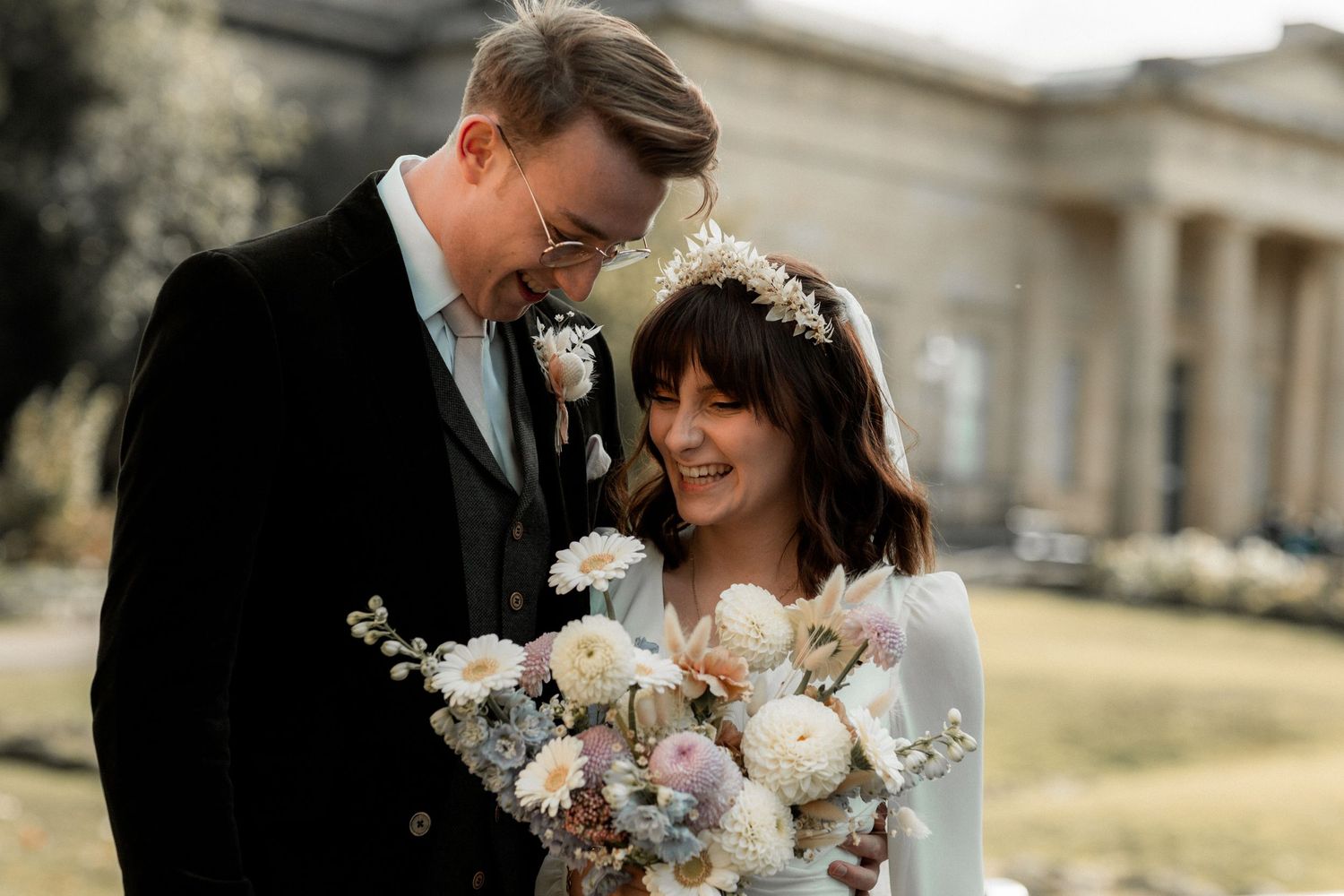 Romantic outdoor wedding portrait with couple sharing laughter and holding delicate floral bouquet.