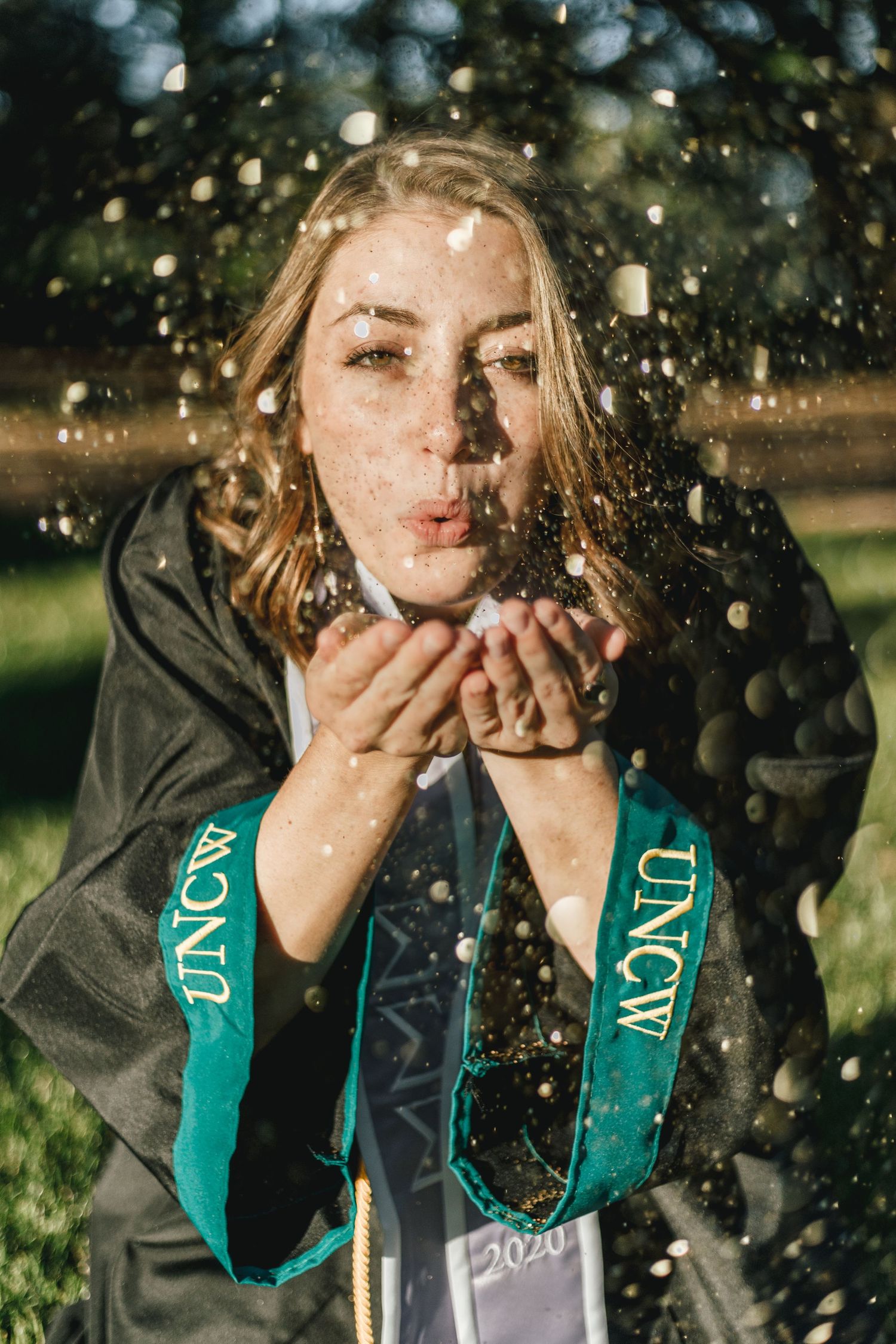 Graduate wearing green graduation stole blows glitter in outdoor celebration photo.