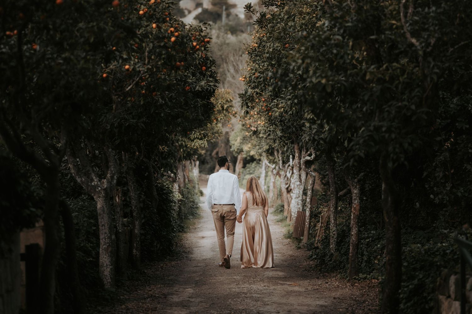 Romantic couple walking down tree-lined path in ethereal outdoor setting.