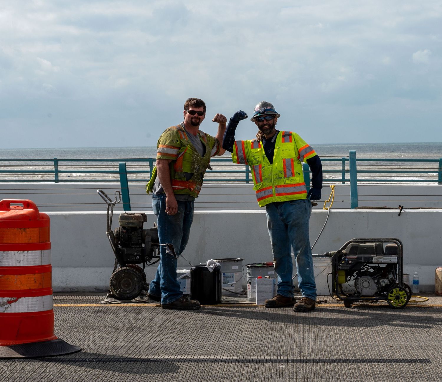 Construction workers in safety vests working on a waterfront project with equipment and orange barriers.