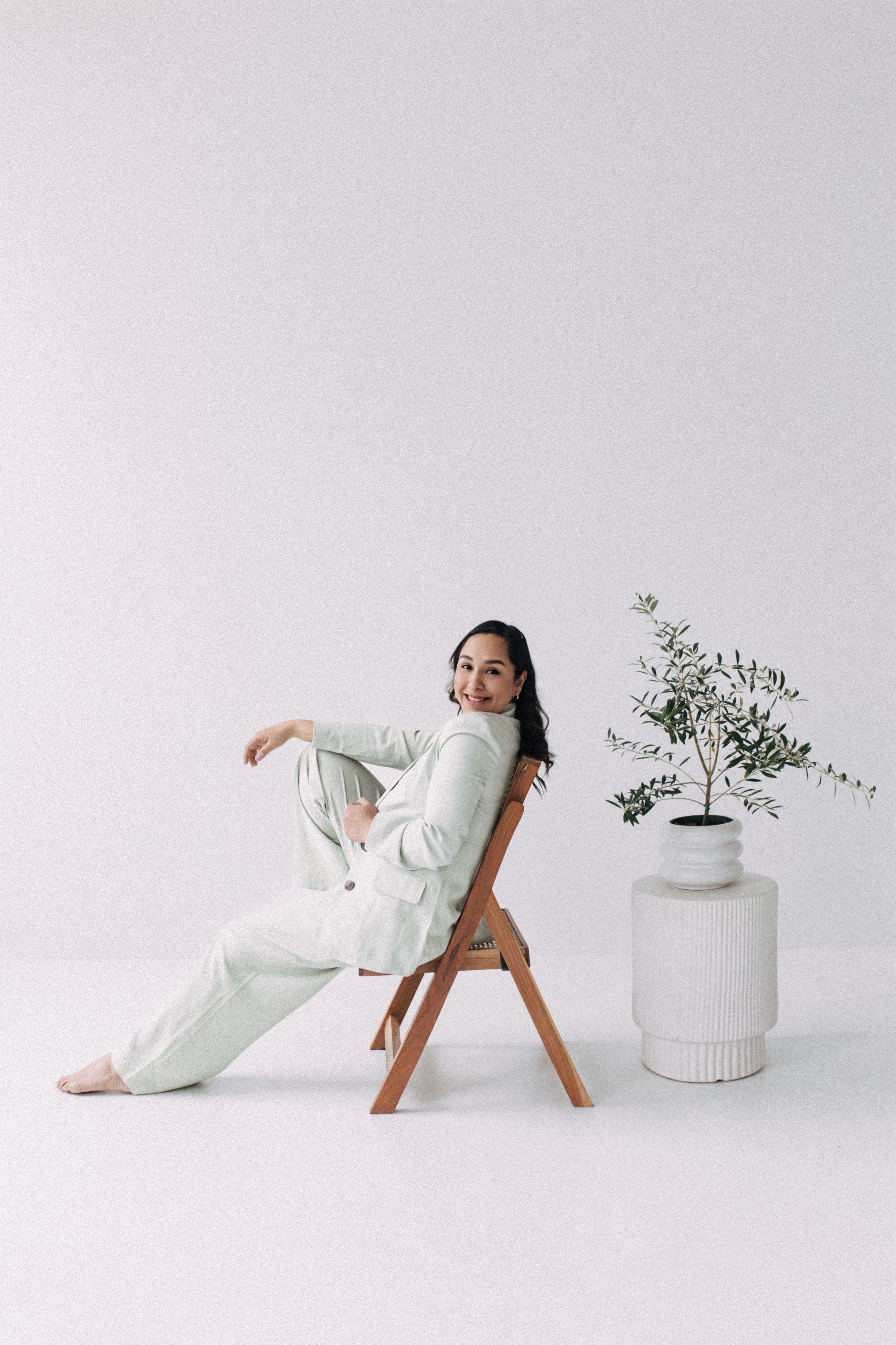 Lifestyle photo of a person in white loungewear posing on wooden chair against white backdrop with plant.