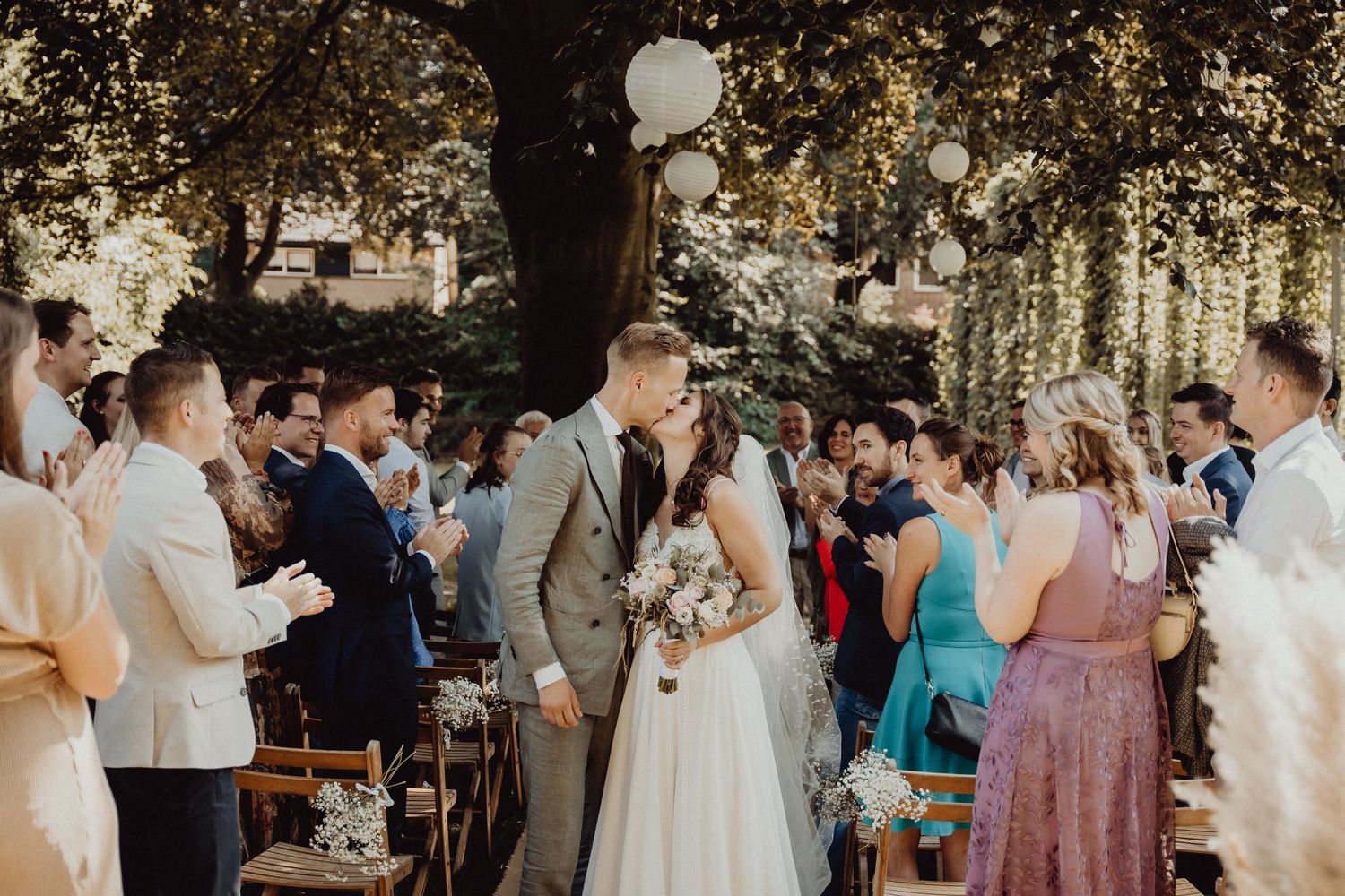 Wedding guests gather under a large tree during an outdoor ceremony.