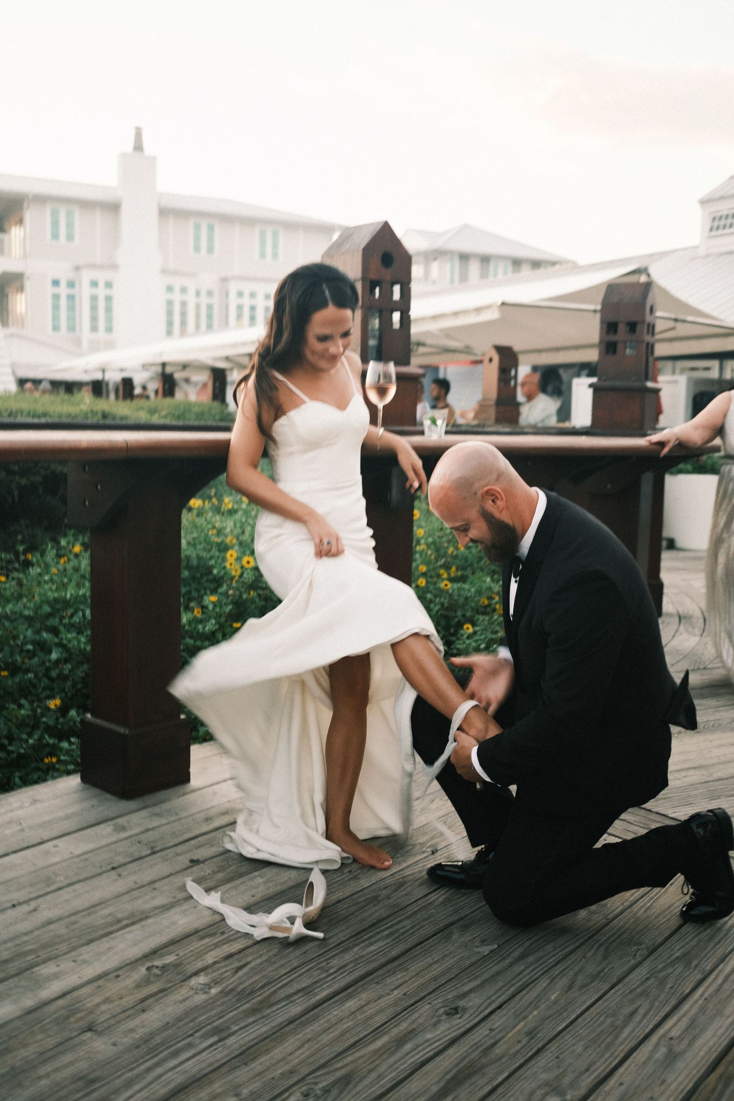 Wedding scene on wooden deck showing dress adjustment and formal attire.