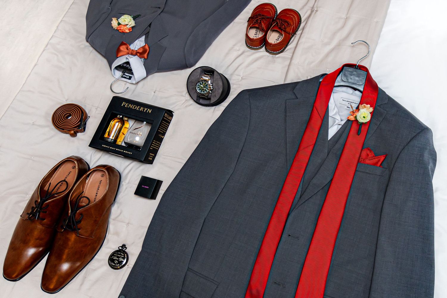 Groom's wedding accessories including grey suit, red tie, brown shoes and cufflinks laid flat.