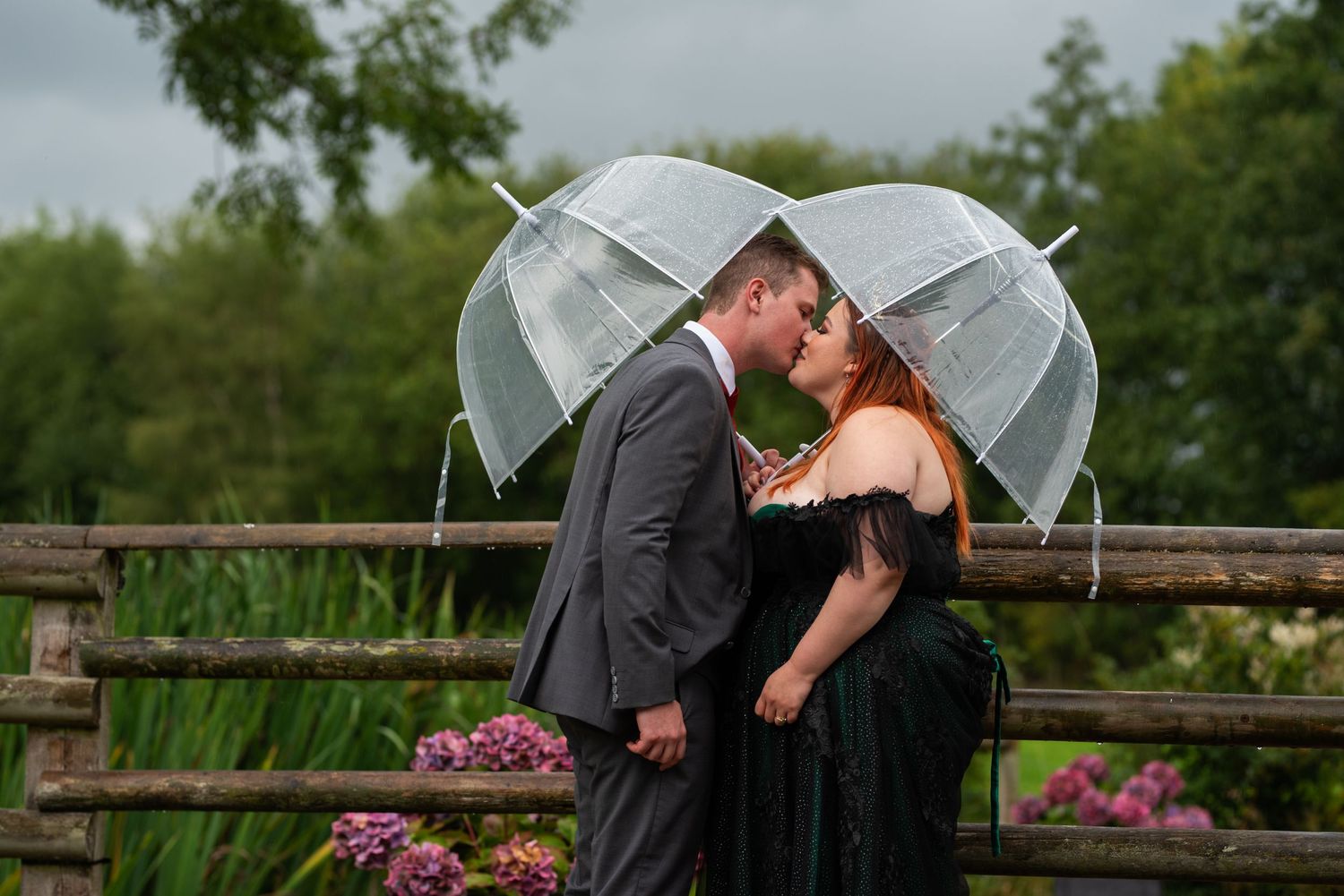 Wedding party members hold clear umbrellas while posing in a garden on a cloudy day.