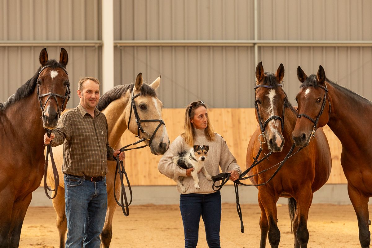 Four horses and their handlers pose together in a stable with a small dog.
