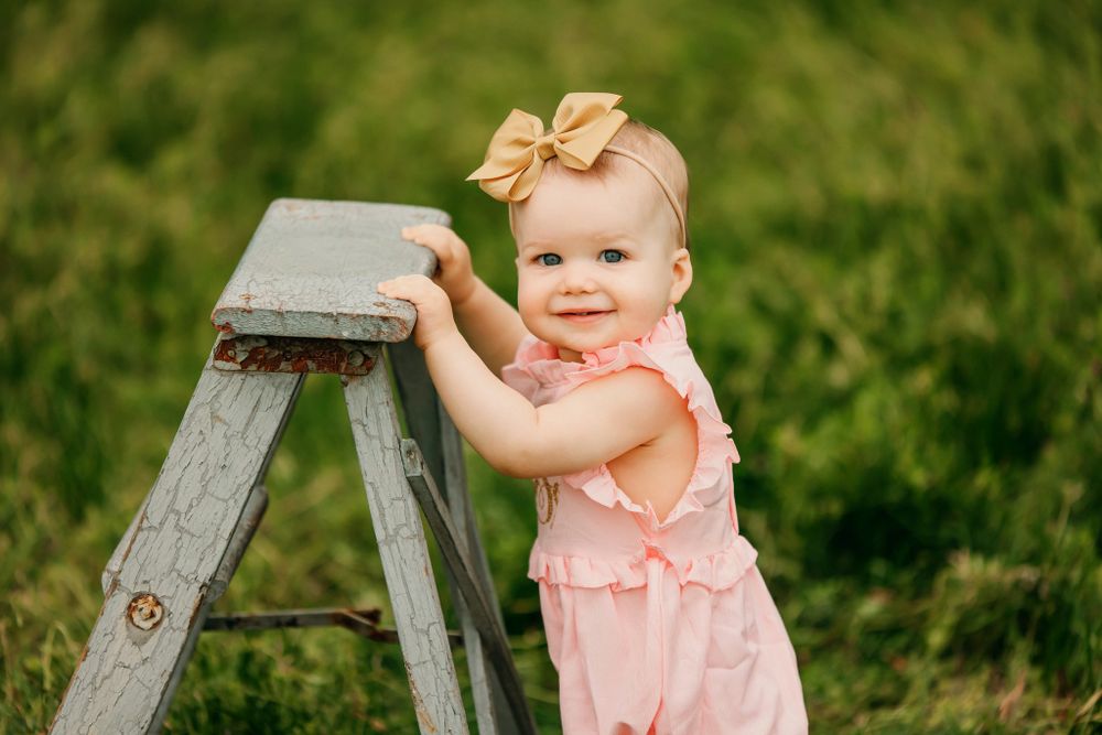 Baby in pink outfit sitting on vintage wooden ladder outdoors.